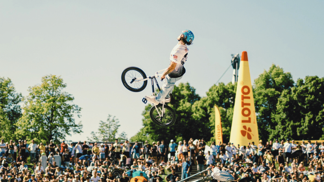 Biker performing a trick in the air at a sports event with a crowd and Lotto branding in the background.
