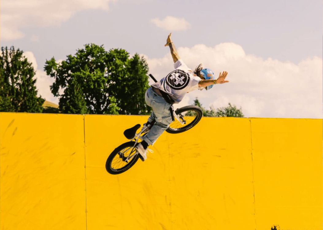 Person on a bicycle performing a trick in mid-air against a yellow wall with 'IOU Ramps' branding.